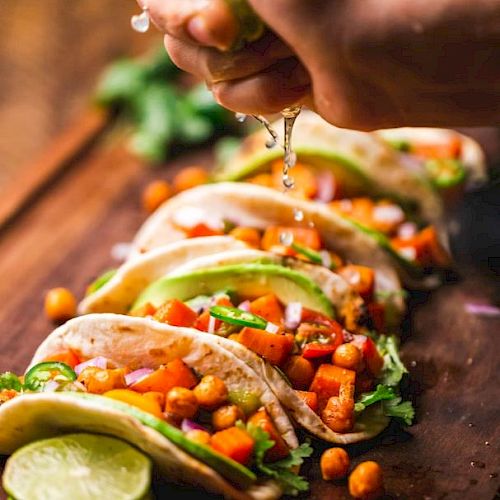 The image shows several tacos filled with chickpeas and vegetables on a wooden board, topped with fresh lime juice being squeezed over them.