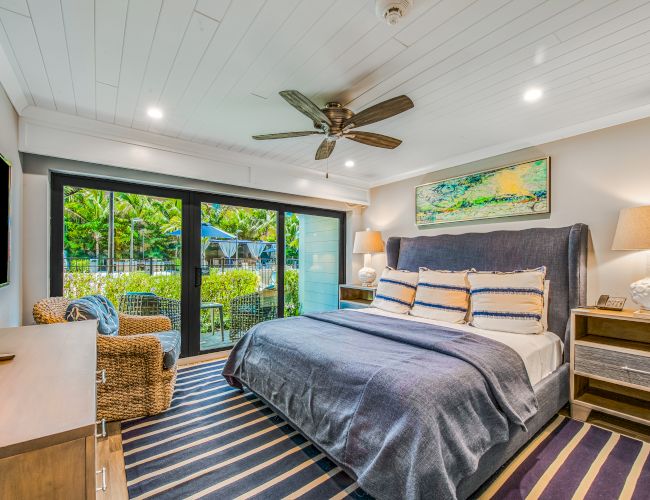 A cozy bedroom featuring a large bed with striped pillows, a wicker chair, a ceiling fan, a TV, and a glass door leading to an outdoor area.