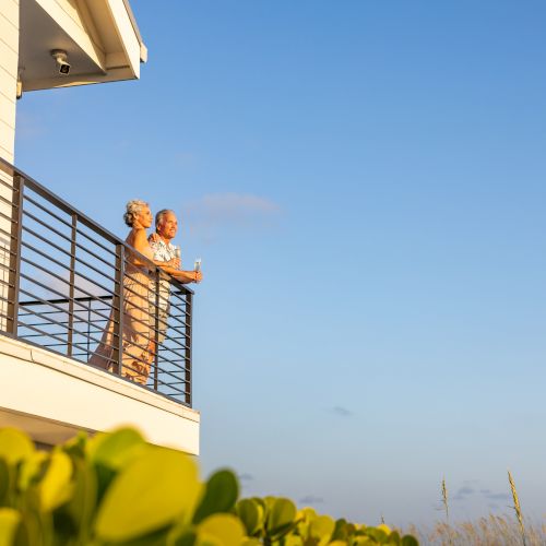 An elderly couple sits on a sunny apartment balcony, sipping drinks and chatting under a clear blue sky.