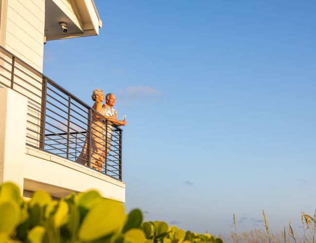 An elderly couple sits on a sunny apartment balcony, sipping drinks and chatting under a clear blue sky.