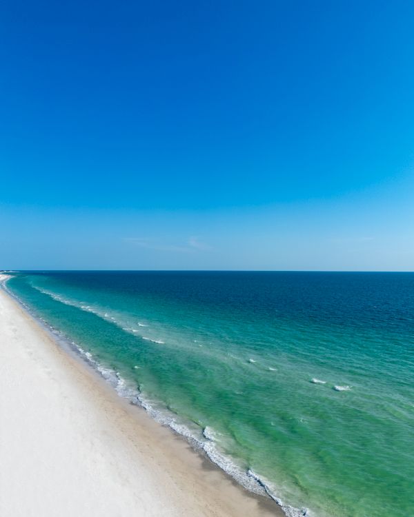 A wide sandy beach stretches along the turquoise ocean with gentle waves under a bright blue sky.