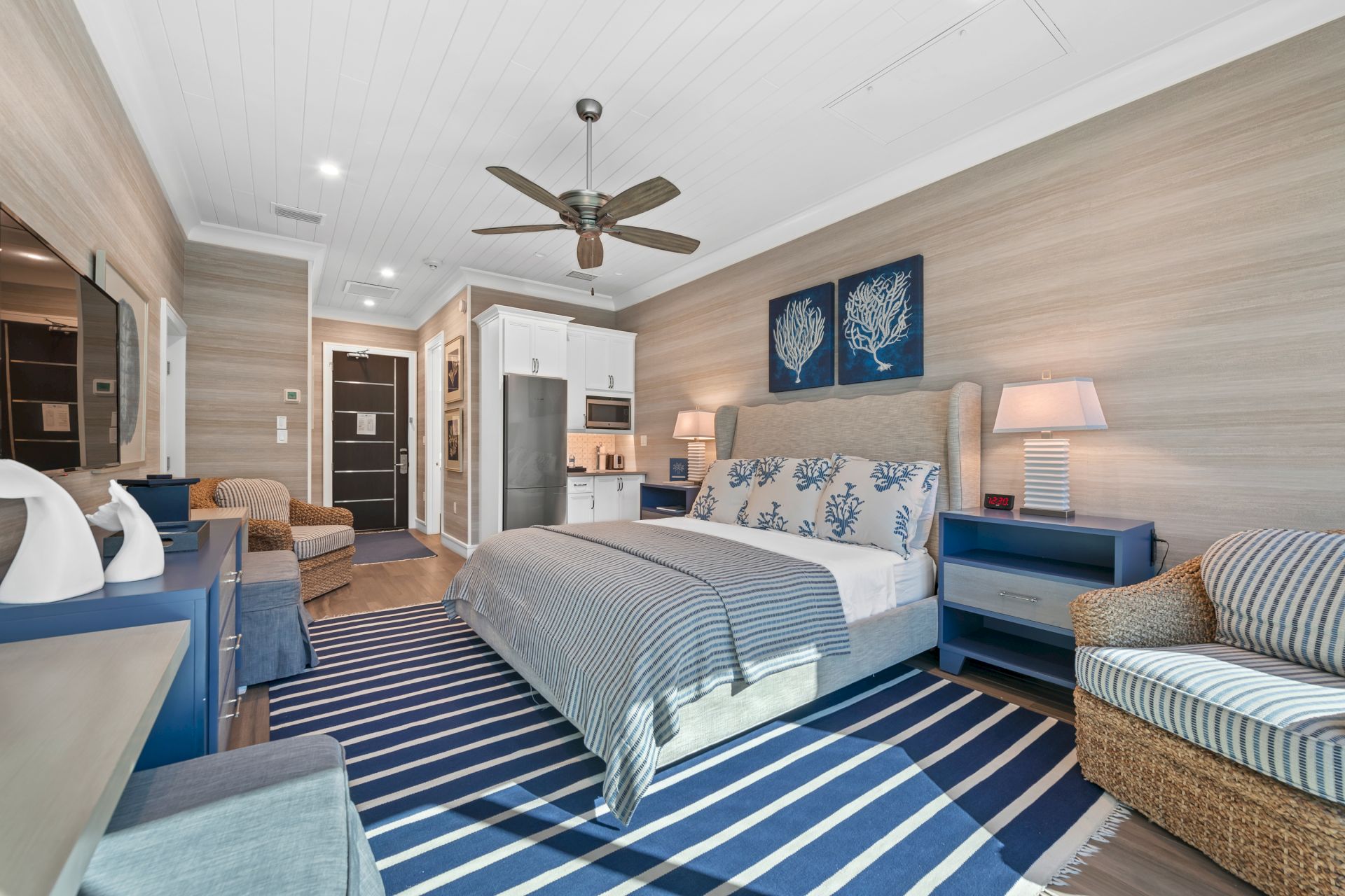 A modern bedroom with a ceiling fan, blue-and-white bedding, nightstands, wall art, and a kitchenette area in the background.