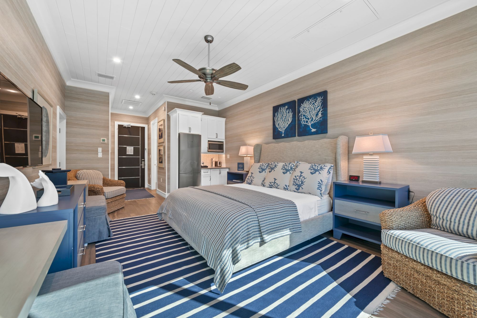 A modern bedroom with a ceiling fan, blue-and-white bedding, nightstands, wall art, and a kitchenette area in the background.