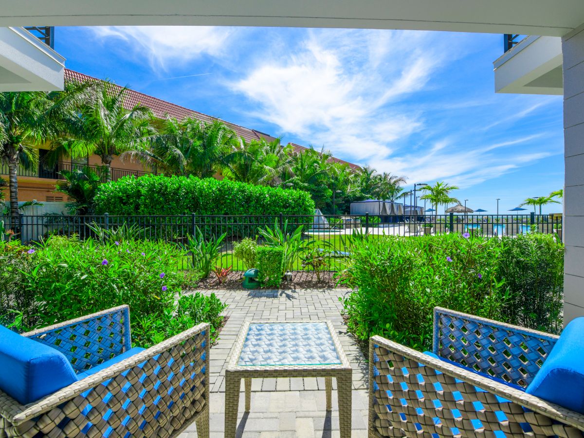 A tropical garden view from a shaded patio with two blue-cushioned wicker chairs and a small table under a vibrant blue sky with scattered clouds.