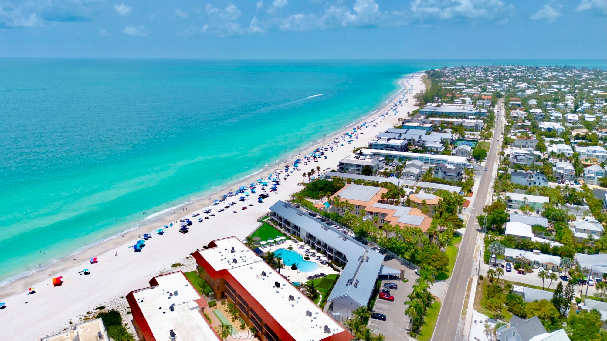 Aerial view of a tropical coastline with turquoise ocean, sandy beach full of umbrellas, and a beachfront hotel by the road.