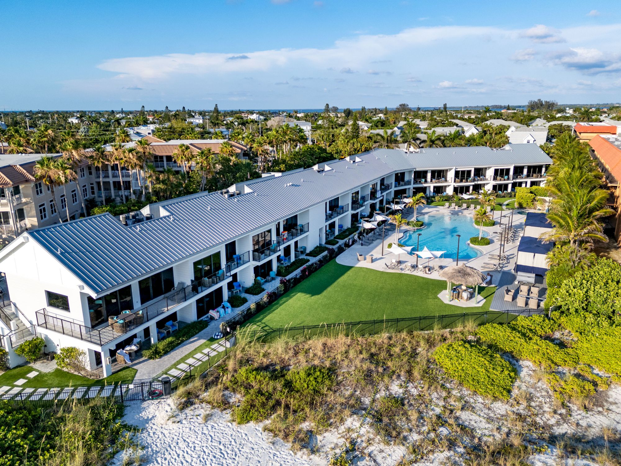 An aerial view of a beachfront resort with a rectangular pool, surrounded by sunbeds, greenery, and adjacent to sandy dunes.