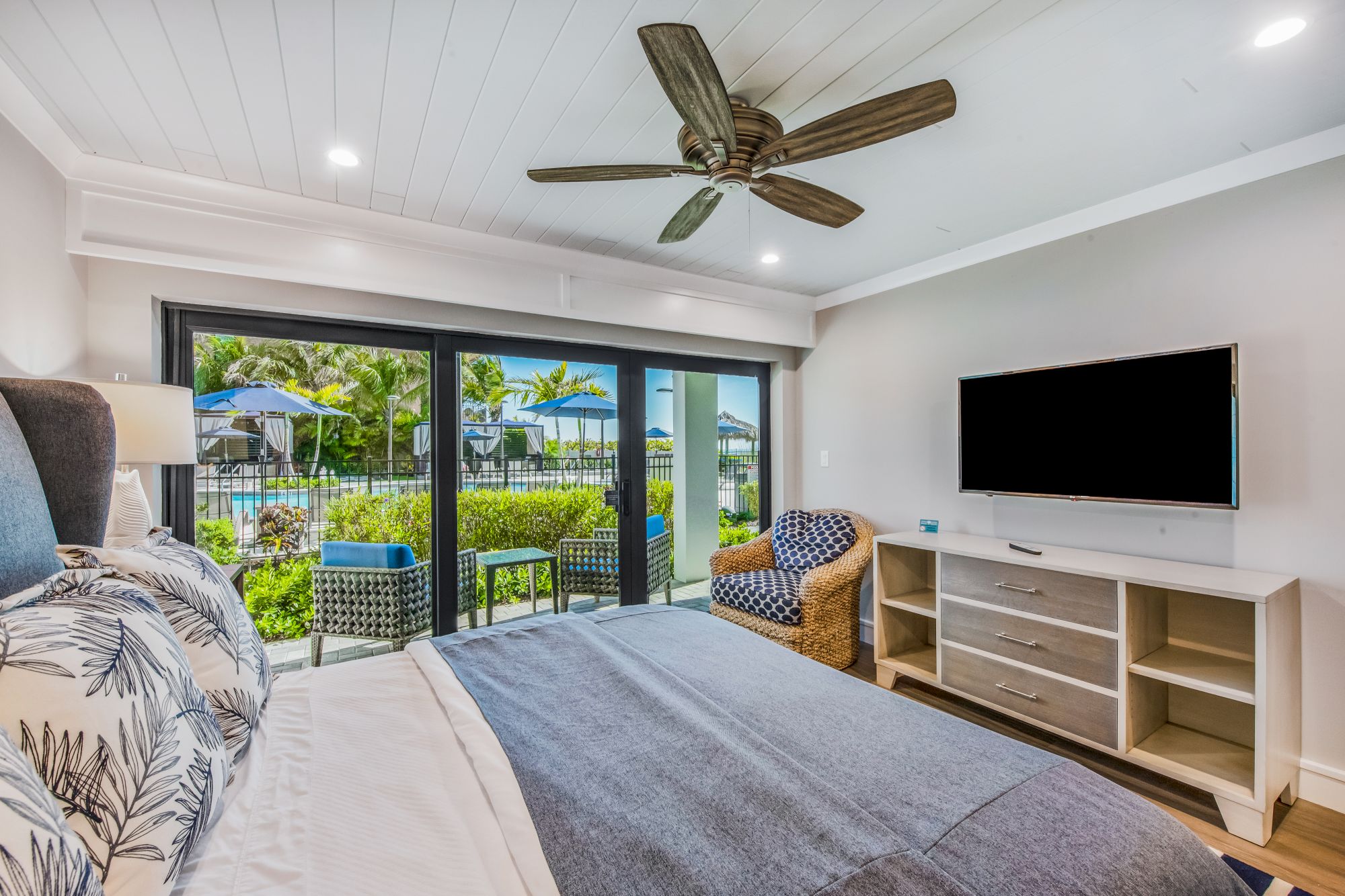 A modern bedroom with a large bed, flat-screen TV, ceiling fan, armchair, and sliding glass doors opening to a patio with outdoor furniture.