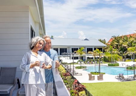 An elderly couple in robes shares coffee on a sunny resort balcony beside a swimming pool and lush gardens.