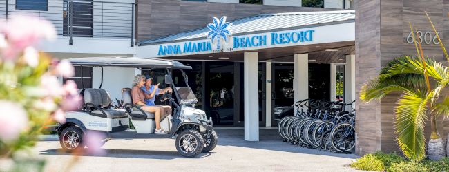 A golf cart with two people is parked in front of Anna Maria Beach Resort, with bicycles and palm trees in the background.