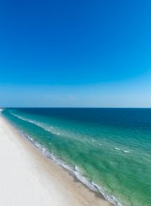 A pristine beach with white sand and clear turquoise water, stretching into the distance under a bright blue sky on a sunny day.