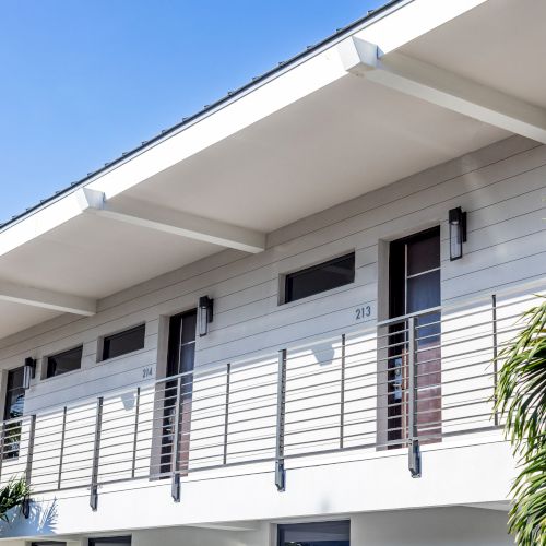 The image shows an exterior view of a modern, white building with a balcony and room numbers, surrounded by palm trees on a sunny day.