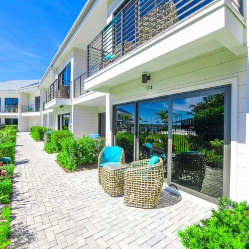 A modern, sunny apartment complex with patio furniture and greenery along a paved walkway. The sky is clear and blue, creating a pleasant atmosphere.