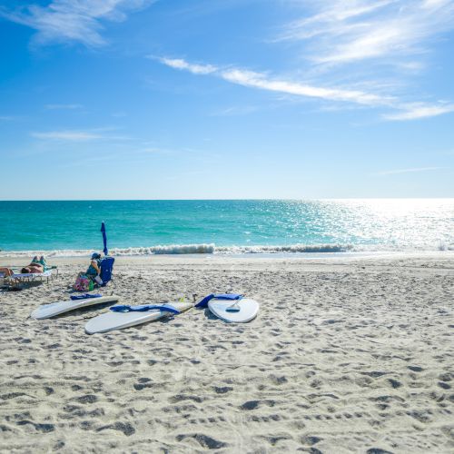 Two people are relaxing on lounge chairs at the beach with paddleboards on the sand, looking out at the calm, blue ocean under a sunny sky.