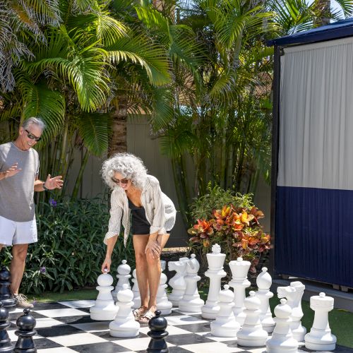 Two people are playing on a large outdoor chessboard with oversized pieces, surrounded by lush greenery.