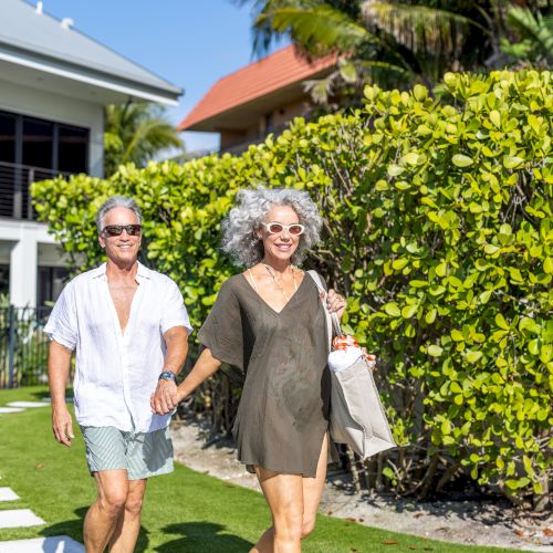 A smiling couple walks hand-in-hand along a garden path on a sunny day, with lush greenery and palm trees in the background, enjoying their stroll.