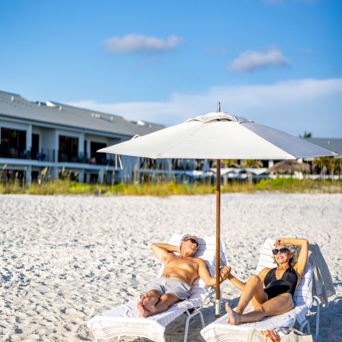 Two people are relaxing on lounge chairs under a beach umbrella on a sandy beach, with buildings visible in the background.