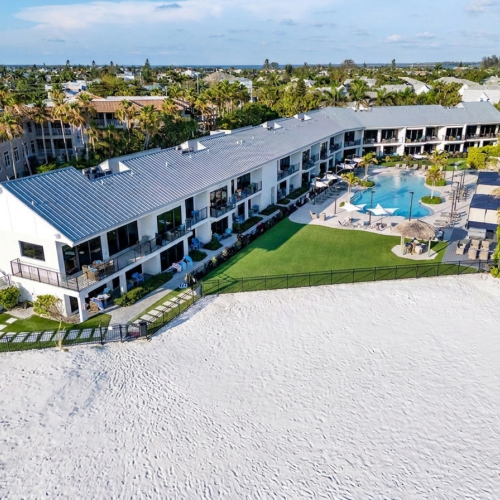 Aerial view of a beachfront resort with modern buildings, a manicured garden, and a swimming pool by the sand.