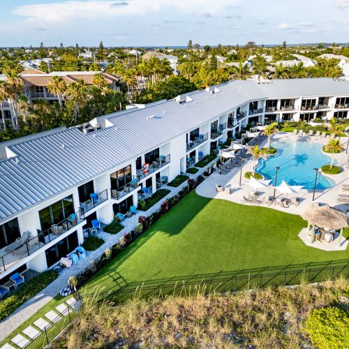 An aerial view of a resort featuring a large pool area, surrounded by loungers and umbrellas, and a large grassy area with adjacent buildings.