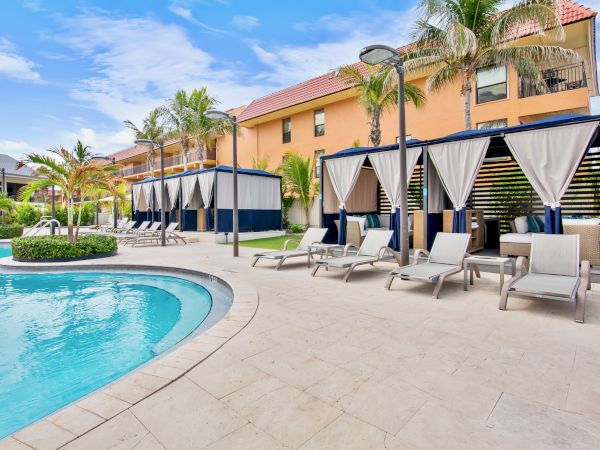 A sunny resort poolside scene with palm trees, lounge chairs under blue-and-white cabanas, and a tiled hotel building in view.