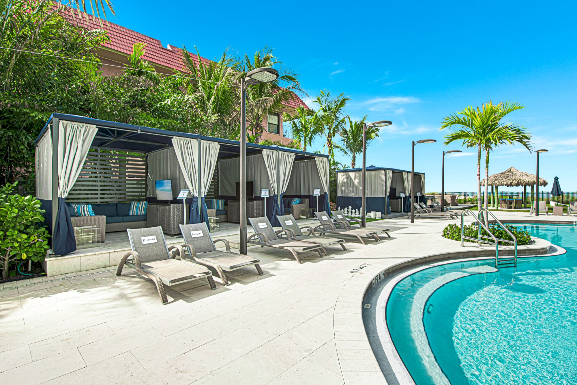 A luxurious poolside area features lounge chairs, cabanas with curtains, palm trees, and a clear blue swimming pool under a bright, sunny sky.