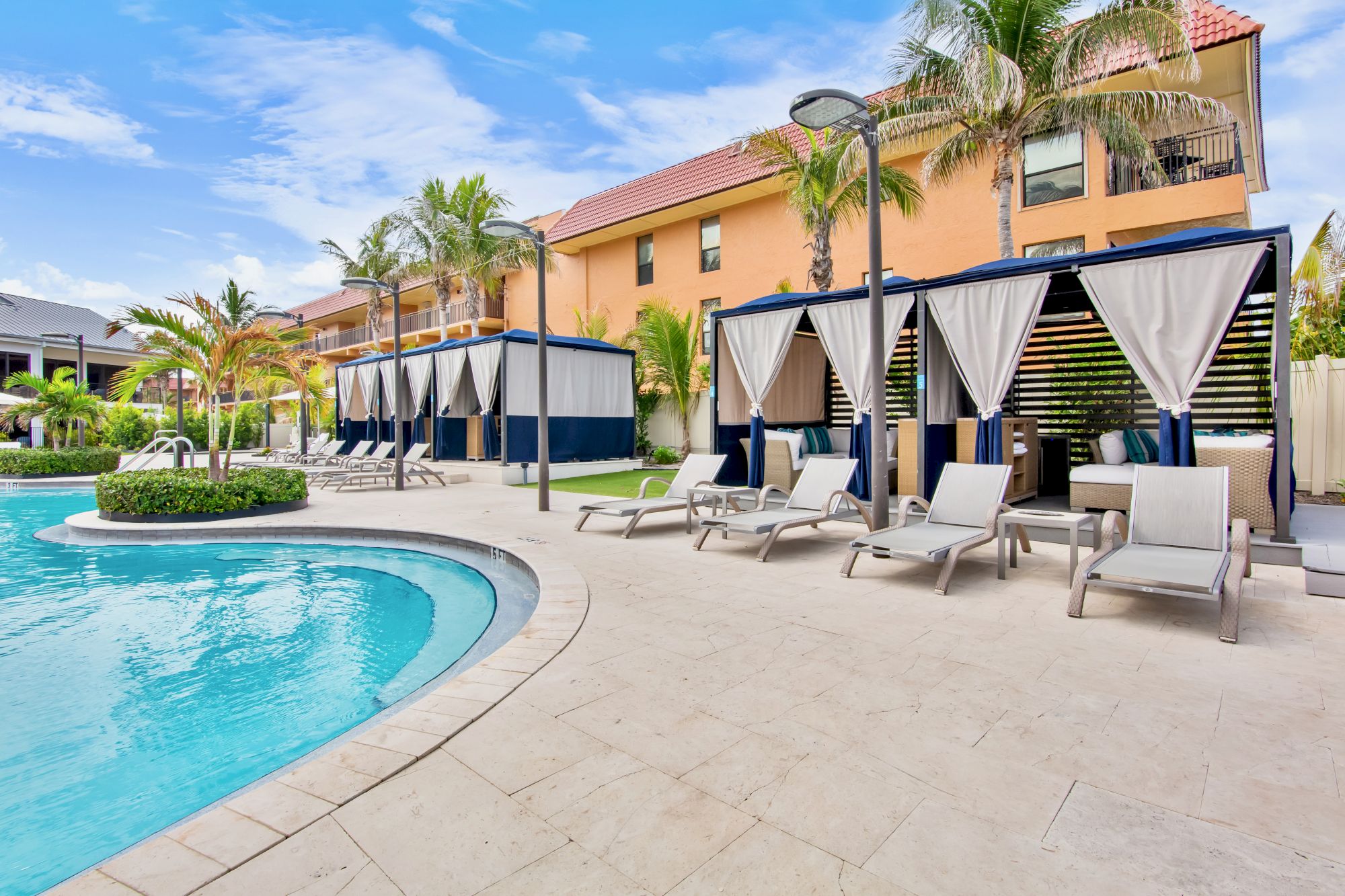 A sunny poolside patio with cabanas, loungers, palm trees, and a yellow building under a bright blue sky.
