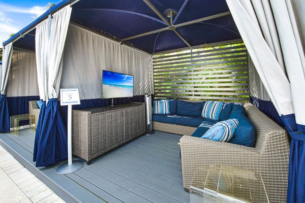 A cozy outdoor cabana with wicker furniture, cushions, a flat-screen TV, curtains, and a small table on a deck.