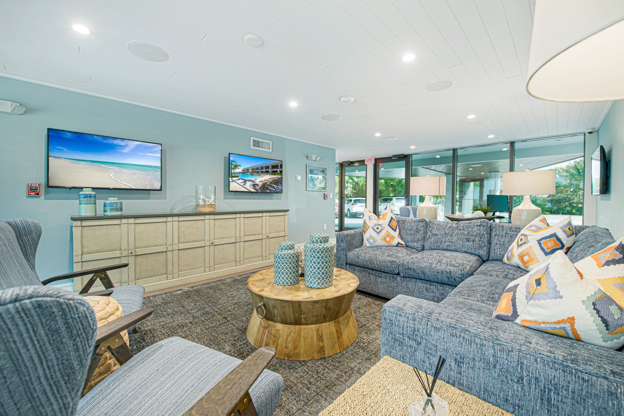 The image shows a modern living room with blue sofas, a wooden coffee table, two wall-mounted TVs, and large windows letting in natural light.