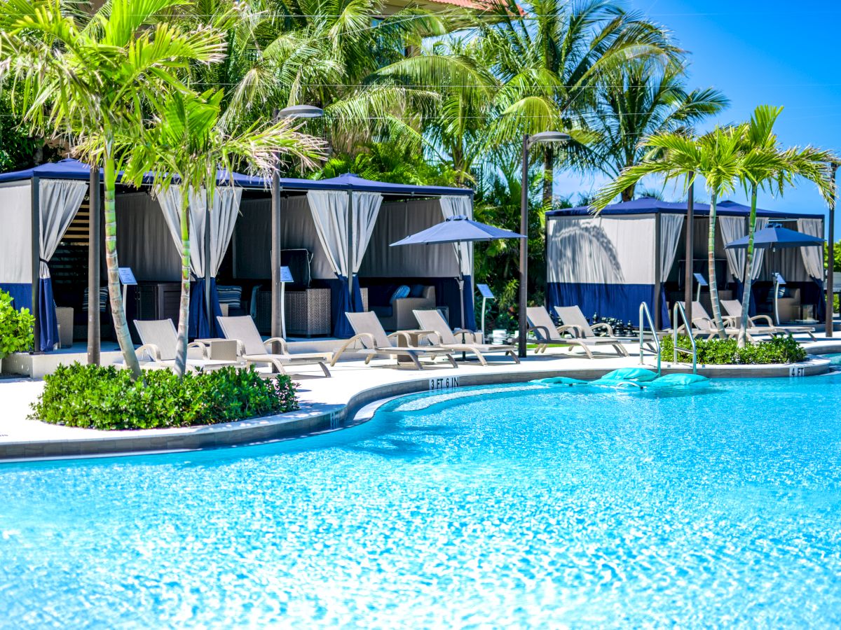 The image shows a luxury pool area with clear blue water, shaded cabanas, lounge chairs, and palm trees in a tropical setting.