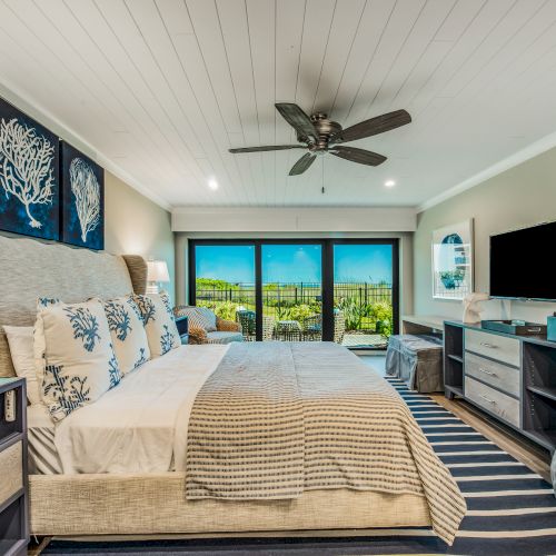 A modern bedroom with a neatly made bed, decorative pillows, ceiling fan, wall art, flat-screen TV, and a view of an outdoor landscape through large windows.