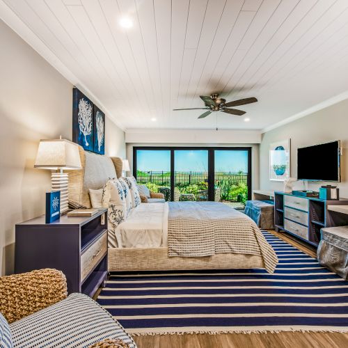 A modern bedroom with a striped rug, bed, armchair, dresser, TV, and large windows overlooking a scenic view, complete with a ceiling fan.