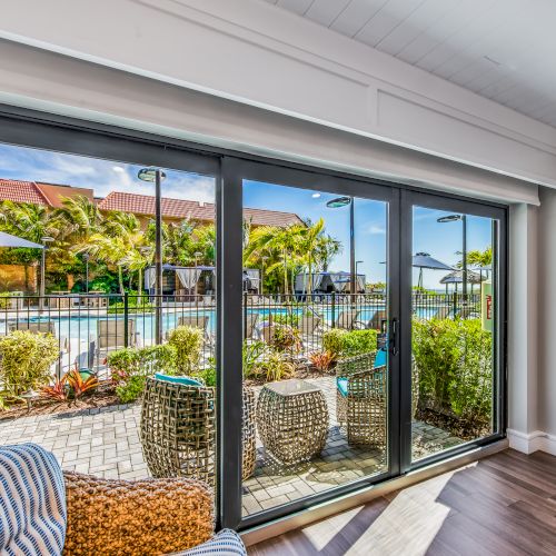 A room with sliding glass doors opens to a patio featuring outdoor seating and a view of a pool surrounded by palm trees and greenery.