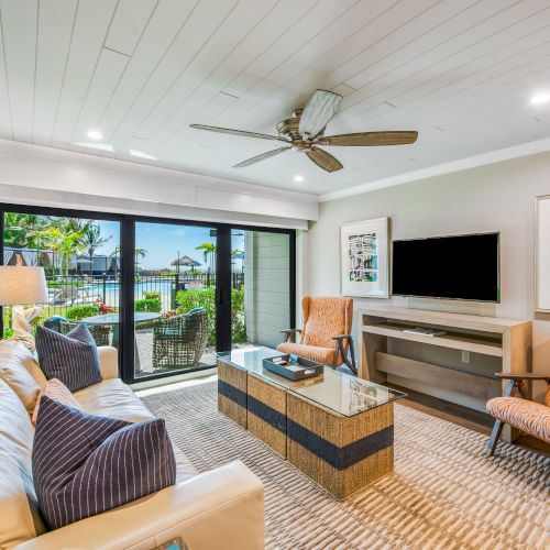 The image shows a modern living room with beige walls, a white ceiling, a ceiling fan, a sofa, two armchairs, a coffee table, and a large sliding glass door.