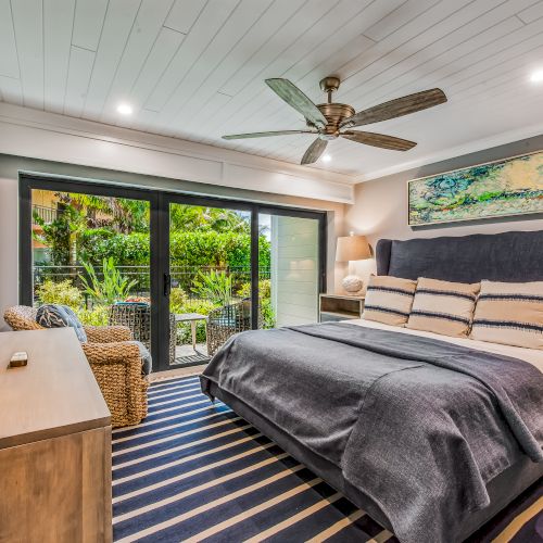 A modern bedroom with a large bed, ceiling fan, striped rug, wall art, dresser, TV, wicker chair, and sliding doors leading to an outdoor view.