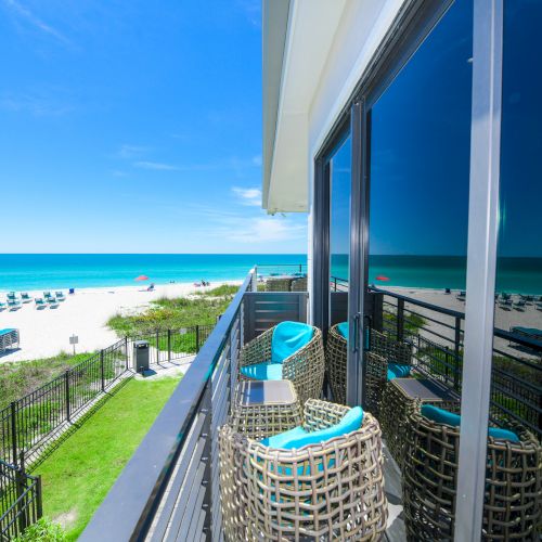 A beachfront balcony with wicker chairs overlooks a pristine sandy beach and blue ocean under a clear sky, reflecting on large windows.