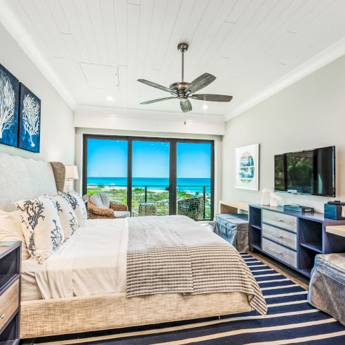 A spacious, modern bedroom with a large bed, flat-screen TV, ceiling fan, art on the walls, and a window offering a view of the ocean can be seen.