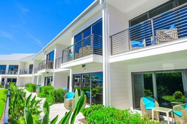 The image shows a modern apartment complex with balconies and patios, green landscaping, and outdoor seating under a clear blue sky.