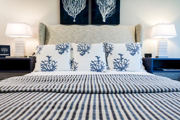 A neatly made bed with striped bedding, three decorative pillows, two lamps on nightstands, and coral artwork on the wall.