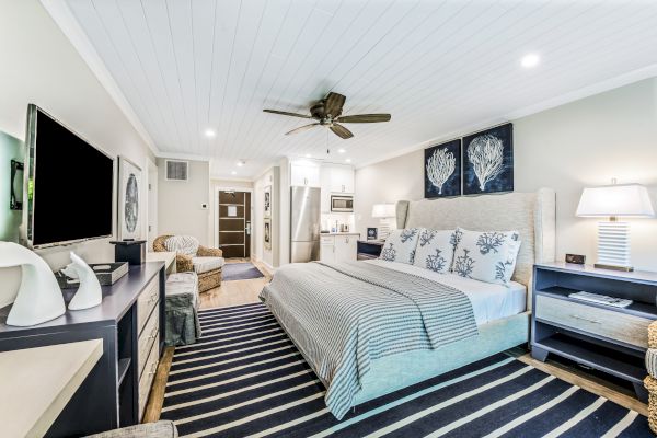 A modern, cozy bedroom features a king-sized bed, striped rug, flat-screen TV, ceiling fan, and light decor with two framed artworks on the wall.