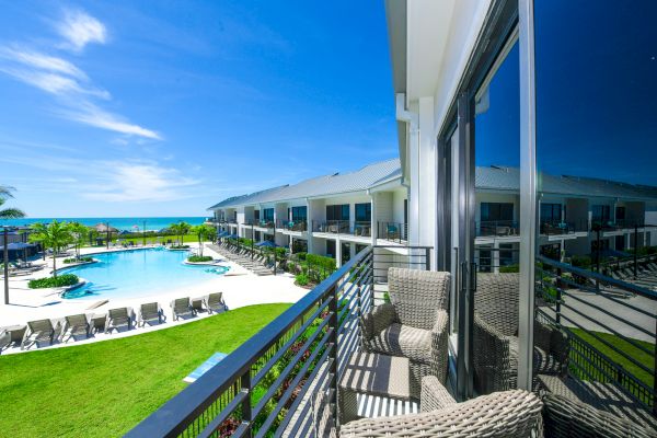 The image shows a balcony overlooking a resort pool area with lounge chairs, palm trees, and a view of the ocean under a clear blue sky.