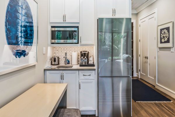 This image shows a small kitchen with a microwave, refrigerator, coffee maker, and cabinets. There is a table to the left with wall art above it.