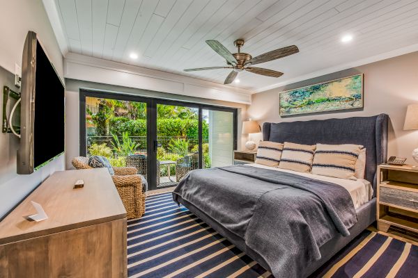 A modern bedroom features a large bed, TV, ceiling fan, wooden furniture, and sliding glass doors opening to a lush garden.