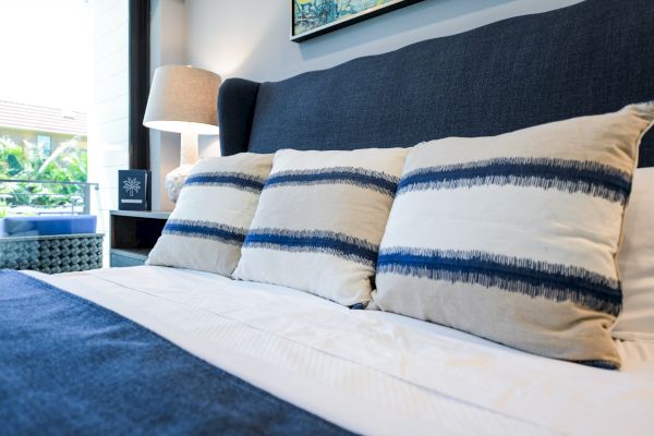 A neatly made bed with blue and beige pillows, a lamp on a bedside table, and a window in the background with a view of greenery.