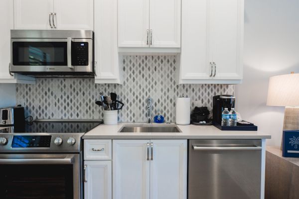 A modern white kitchen with stainless steel appliances, a sink, counter with utensils, coffee maker, and a lamp on the side, and a tiled backsplash.