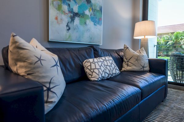 A modern living room with a dark blue sofa decorated with patterned pillows, an abstract painting, a floor lamp, and a view of a balcony outside.