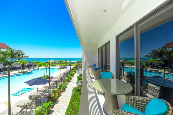 The image shows a luxurious balcony with seating overlooking a beautiful pool area and the ocean, featuring palm trees and clear blue skies.