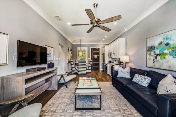 A modern living room features a ceiling fan, wall-mounted TV, comfortable seating, a coffee table, and open access to a kitchen area.