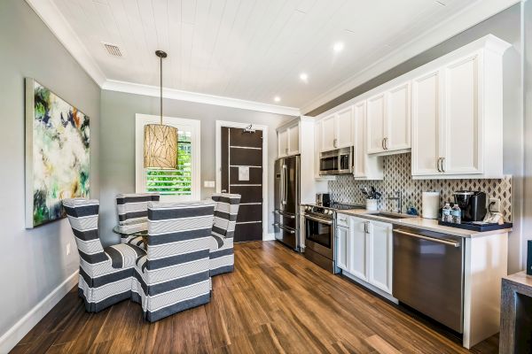 A modern kitchen with wood flooring, white cabinets, stainless steel appliances, a dining table with striped chairs, and a wall painting is shown.