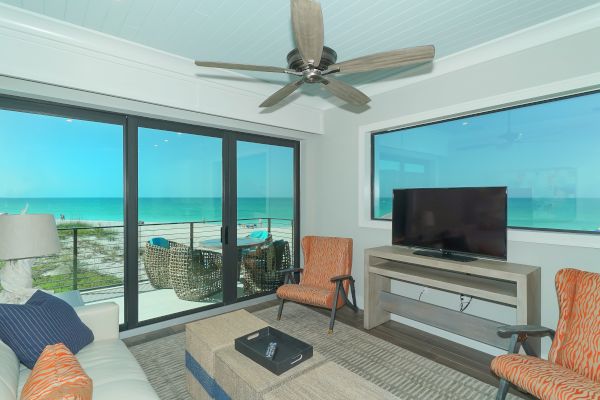 A living room with beach view features a ceiling fan, TV, two chairs, coffee table, large window, and sliding glass doors to a balcony ending the sentence.