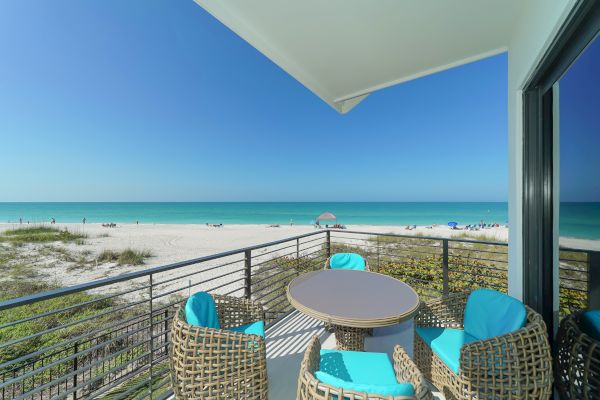 A beachside balcony features rattan chairs with blue cushions around a table, overlooking a sandy beach and clear blue sea under a bright sky.