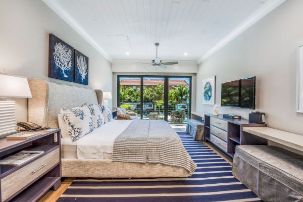 A modern bedroom features a large bed with decorative pillows, a TV, a desk, and glass doors leading to a balcony with outdoor scenery.